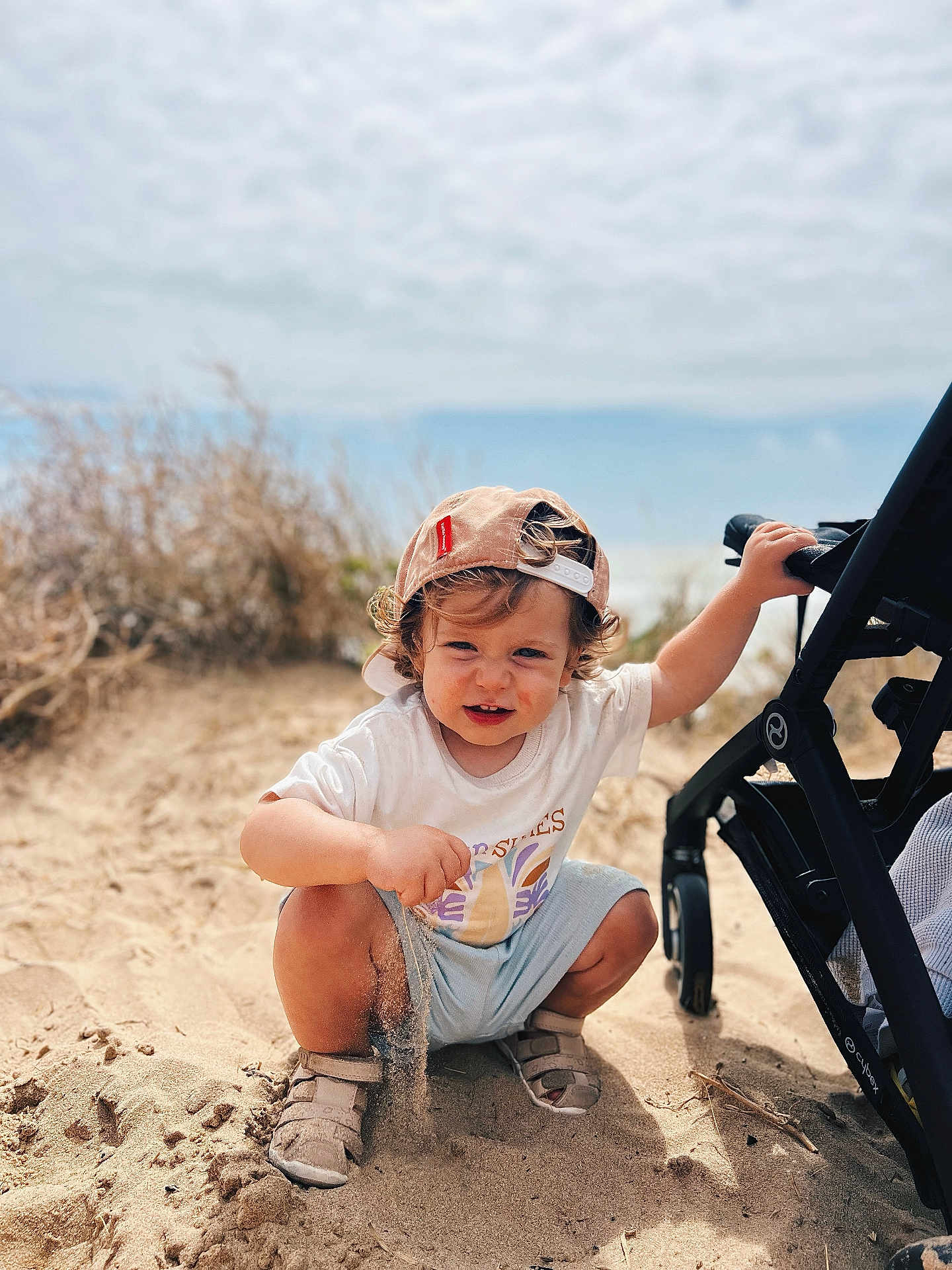 Roméo participe au concours pour gagner de l'argent avec cette photo : beach, cap, child, curly_hair, daylight, footwear, grass, nature, outdoor, person, playing, sand, shorts, sky, smiling, squatting, stroller, sunny, toddler, tshirt