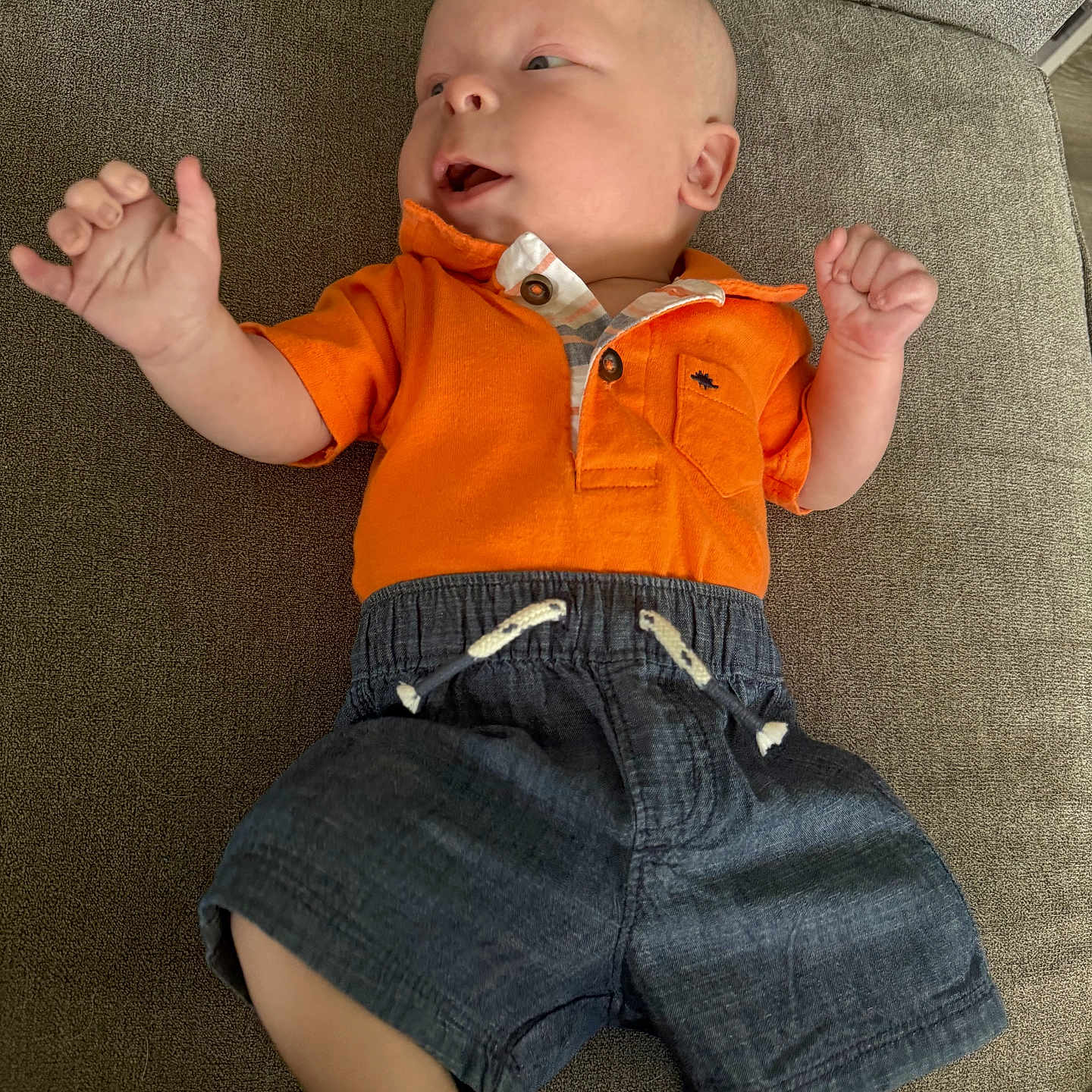 Paxtyn is registered to the contest to win money with this photo: baby, infant, child, orange_shirt, shorts, couch, furniture, smiling, happy, hands, feet, portrait, indoor, relaxing, clothing, person, skin, head, face, laying