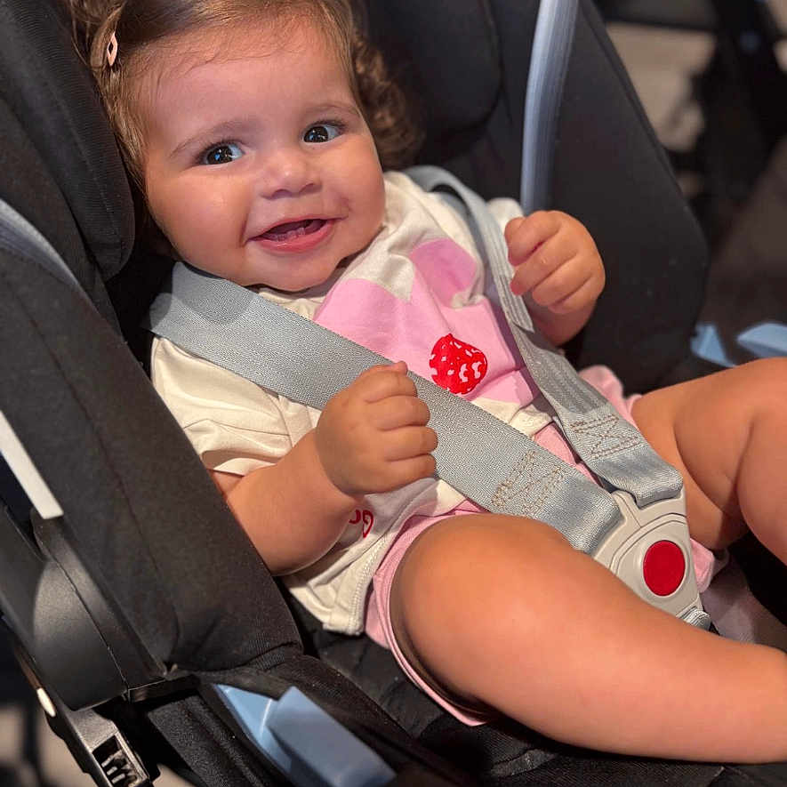 Nour is registered to the contest to win money with this photo: baby, car_seat, cheeks, child, closeup, clothing, curly_hair, cute, happy, harness, indoor, infant, person, pink_clip, portrait, safety, seatbelt, sitting, smile, toddler