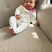 Nour participe au concours pour gagner de l'argent avec cette photo : baby, infant, smiling, white_pajamas, flower_hair_clip, couch, beige_couch, textured_fabric, indoor, natural_light, happy, child, person, cute, cozy, relaxed, portrait, small_hands, feet, home
