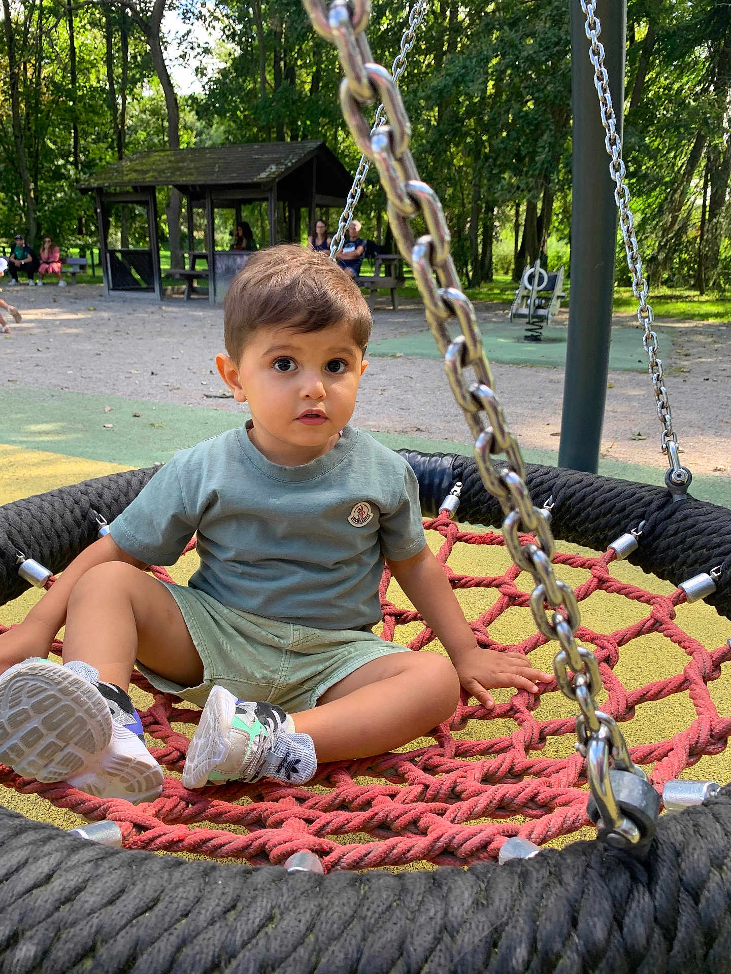 Youyou participe au concours pour gagner de l'argent avec cette photo : child, toddler, swing, playground, outdoor, trees, rope, seat, park, greenery, casual_clothing, shorts, tshirt, sneakers, daylight, people_in_background, bench, recreation, nature, curious_expression