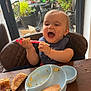 baby, high_chair, spoon, food, messy, plate, bread, table, window, glass_door, garden, car, plant, indoor, child, happy, smiling, feeding, blue_plate, red_spoon