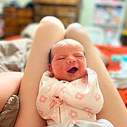 Oaklynn is registered to the contest to win money with this photo: baby, newborn, infant, person, legs, bedroom, blanket, clothing, happy, smiling, face, skin, swaddle, pattern, flower, indoor, soft_focus, closeup, cute, resting