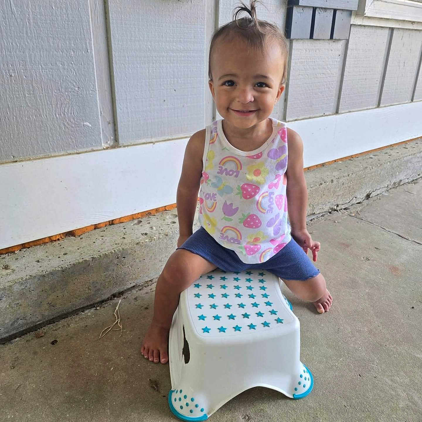 Renli is registered to the contest to win money with this photo: barefoot, blue, child, concrete, cute, daylight, hair_tie, happy, outdoor, person, portrait, shorts, sitting, smiling, stars, stool, tank_top, toddler, wall, white