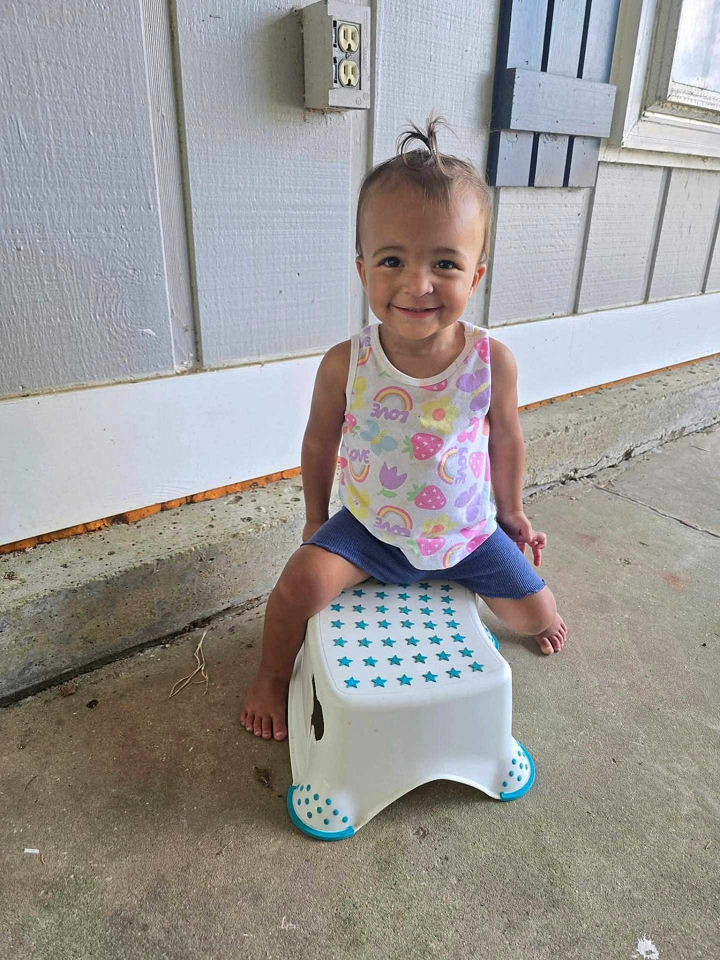 Renli is registered to the contest to win money with this photo: toddler, child, smiling, sitting, stool, white, blue, stars, concrete, wall, outdoor, barefoot, shorts, tank_top, hair_tie, happy, cute, person, portrait, daylight