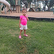 Khori is registered to the contest to win money with this photo: child, playground, grass, slide, climbing_structure, tree, pink_shirt, shorts, outdoor, daylight, person, footwear, play_equipment, park, casual_clothing, young_child, curious_expression, summer, recreation, nature