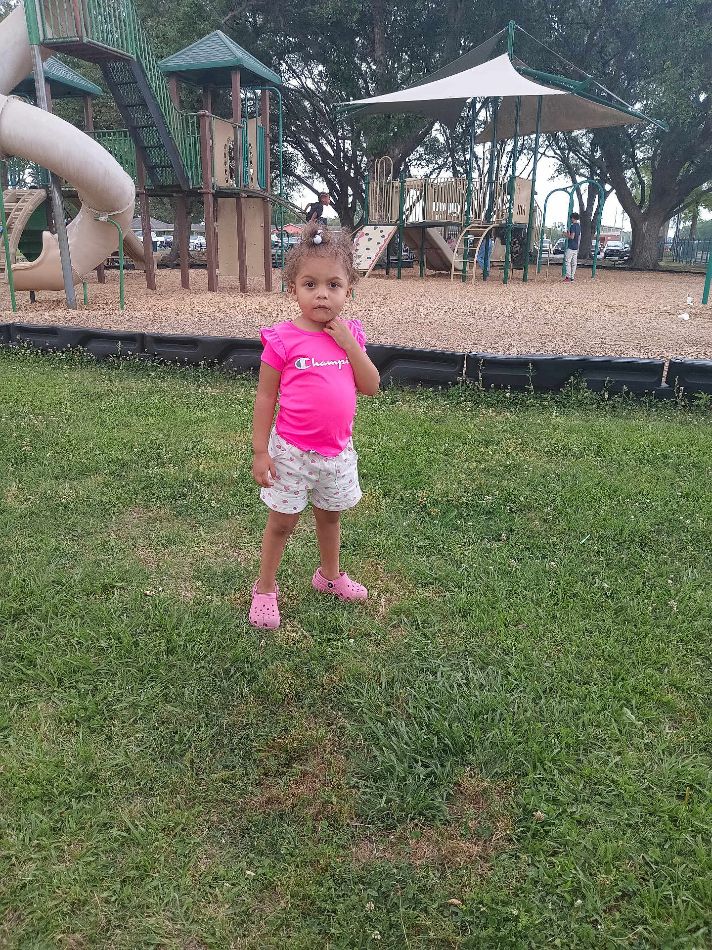 Khori is registered to the contest to win money with this photo: child, playground, grass, slide, climbing_structure, tree, pink_shirt, shorts, outdoor, daylight, person, footwear, play_equipment, park, casual_clothing, young_child, curious_expression, summer, recreation, nature
