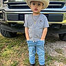 child, boy, cowboy_hat, jeans, tshirt, boots, necklace, cross, truck, ford, outdoor, grass, dirt, vehicle, portrait, standing, casual_clothing, young_child, hands_in_pockets, daytime
