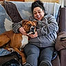 black_bag, blanket, brown_dog, casual, comfort, couch, dog, domestic, furniture, gray_hoodie, happy, human_animal_bond, indoor, living_room, papers, pet, relaxed, seated, smile, woman