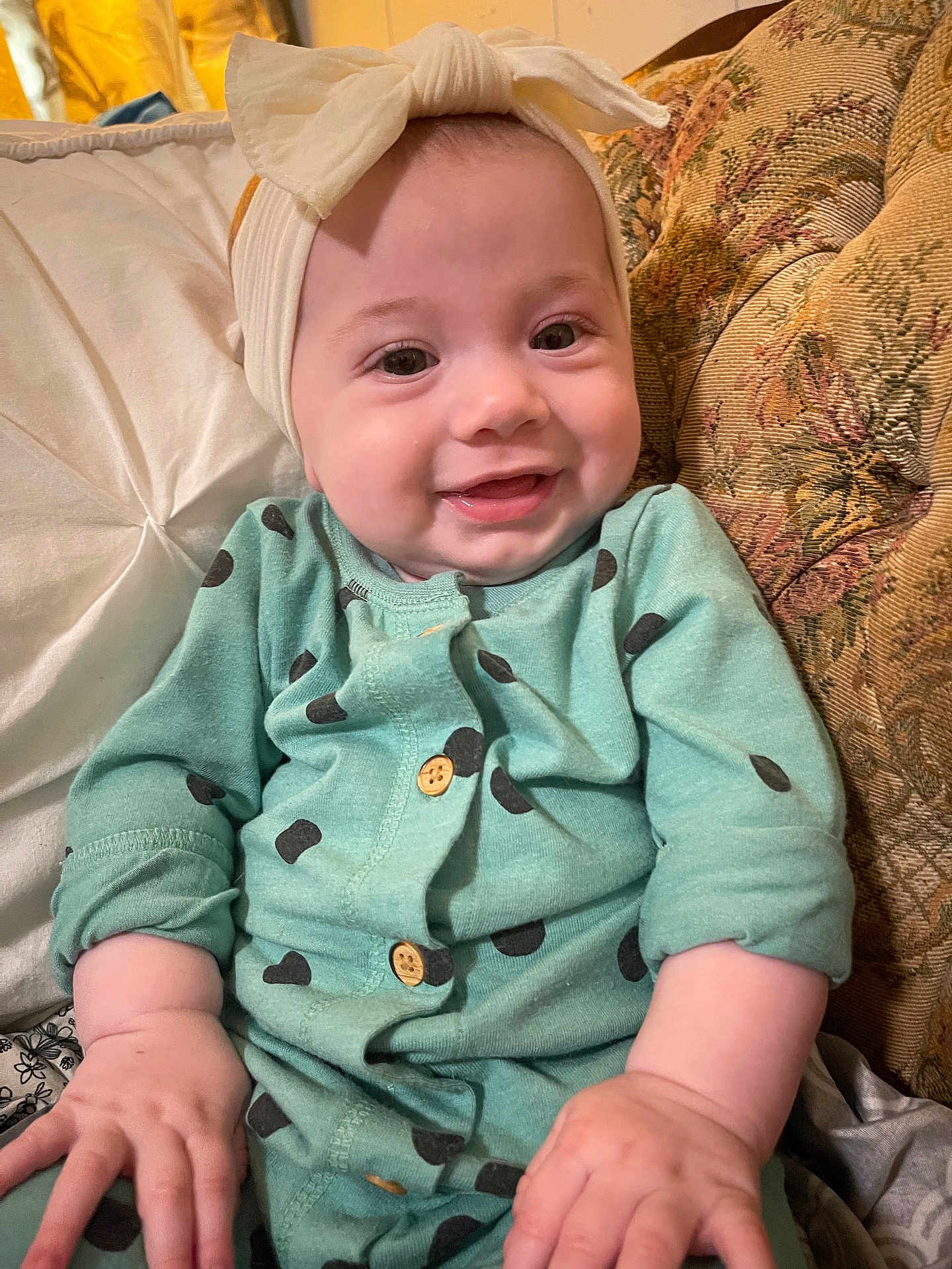 Lydia Faye Anne is registered to the contest to win money with this photo: baby, child, smiling, headband, onesie, polka_dots, couch, pillow, indoor, infant, clothing, cute, happy, portrait, sitting, hand, face, person, fabric, cozy