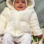 Minha Umer is registered to the contest to win money with this photo: baby, child, smiling, white_jacket, hood, bear_ears, blanket, patterned_blanket, white_pants, cozy, cute, infant, lying_down, happy, indoor, soft_texture, warm_clothing, portrait, person, toddler