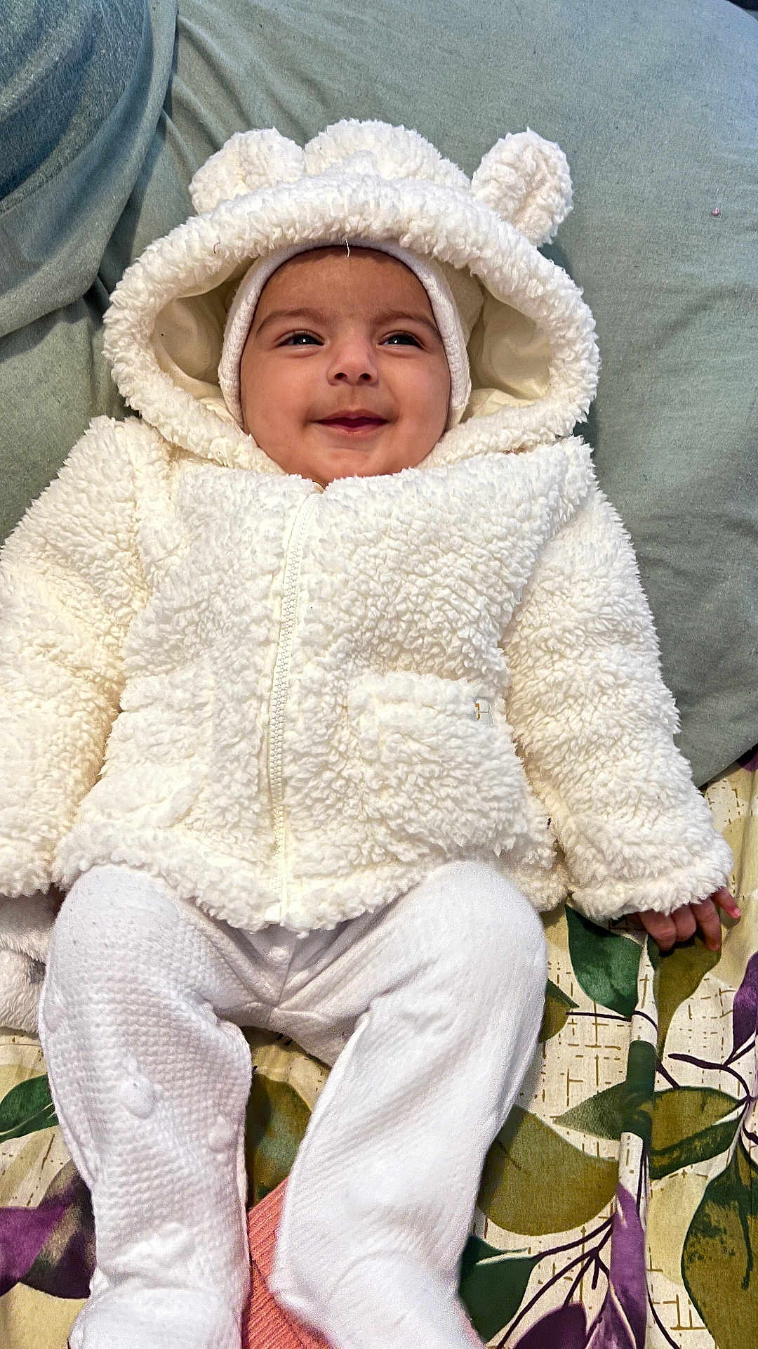 Minha Umer is registered to the contest to win money with this photo: baby, child, smiling, white_jacket, hood, bear_ears, blanket, patterned_blanket, white_pants, cozy, cute, infant, lying_down, happy, indoor, soft_texture, warm_clothing, portrait, person, toddler