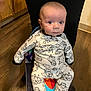 baby, child, onesie, sheep_pattern, chair, wooden_floor, person, indoor, footwear, sitting, curious, wide_eyes, infant, cute, sleepwear, colorful, black_pants, flooring, young_child, portrait