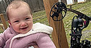 Brinley is registered to the contest to win money with this photo: archery, baby, blanket, child, cold_weather, compound_bow, forest, grass, happy, holding, knitwear, nature, outdoor, person, pink_jacket, recreation, smiling, target, trees, wooden_post