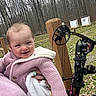 Brinley is registered to the contest to win money with this photo: baby, child, smiling, pink_jacket, blanket, outdoor, forest, trees, compound_bow, archery, target, wooden_post, grass, person, holding, cold_weather, knitwear, happy, nature, recreation