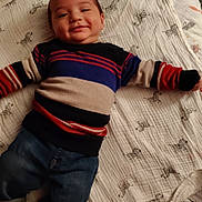 Jonathan is registered to the contest to win money with this photo: baby, child, smiling, striped_sweater, denim_pants, blanket, zebra_pattern, lying_down, happy, indoor, cute, infant, clothing, person, soft_texture, cozy, casual_wear, head, face, toddler