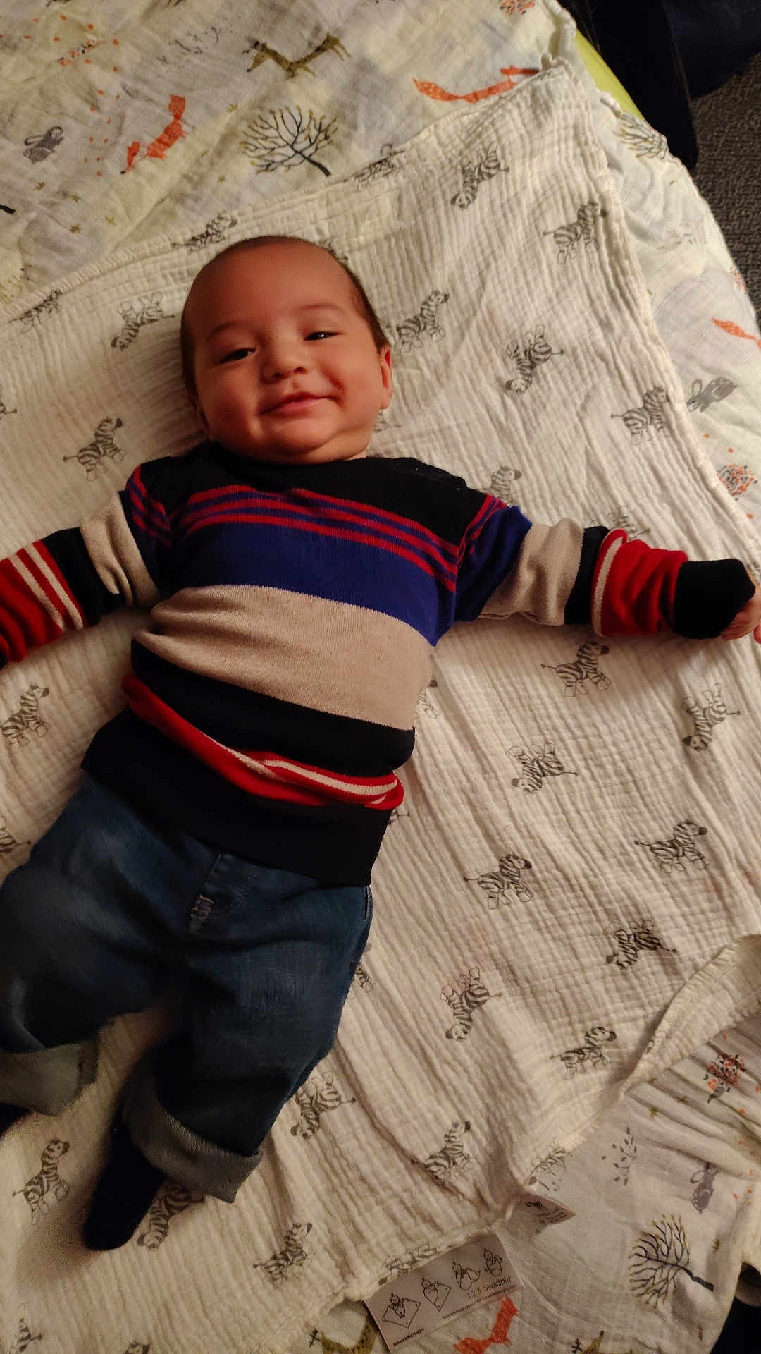 Jonathan is registered to the contest to win money with this photo: baby, child, smiling, striped_sweater, denim_pants, blanket, zebra_pattern, lying_down, happy, indoor, cute, infant, clothing, person, soft_texture, cozy, casual_wear, head, face, toddler
