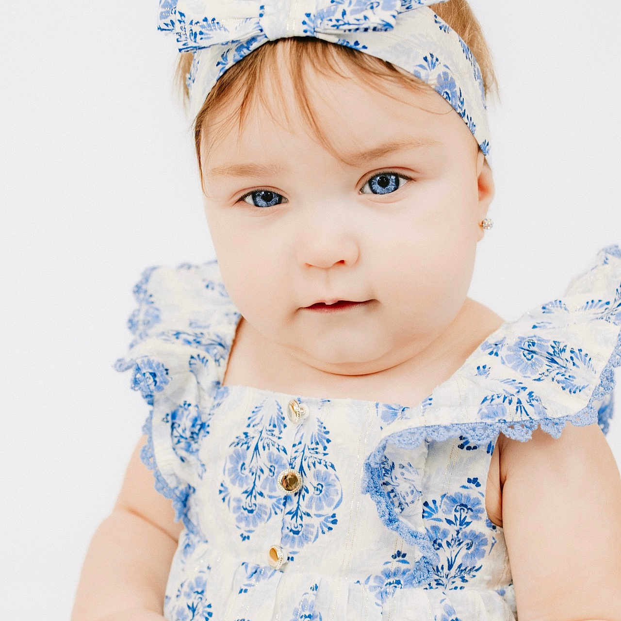 Skyla is registered to the contest to win money with this photo: baby, child, girl, headband, blue_eyes, dress, floral_pattern, cute, portrait, infant, fashion, white_background, soft_lighting, closeup, hands, face, eyewear, earrings, smile, innocence