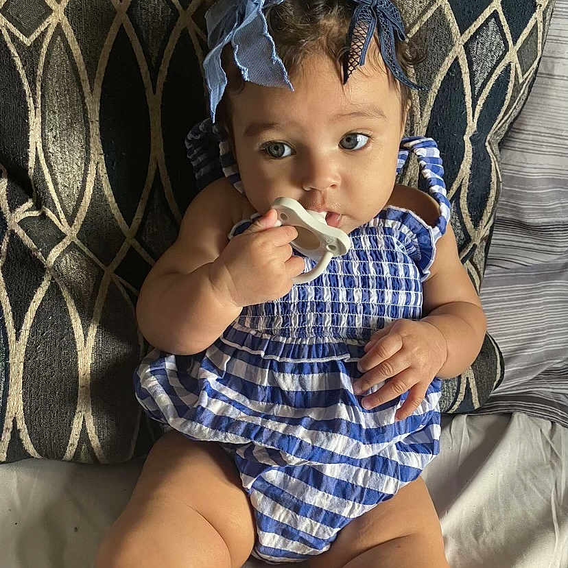 Jahnova is registered to the contest to win money with this photo: baby, blue_bow, child, chubby, comfort, curly_hair, cute, eyes, face, hand, indoors, infant, legs, pacifier, patterned_background, pillow, portrait, sitting, skin, striped_outfit