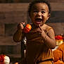 toddler, child, pumpkin, autumn, fall, orange, brown_clothing, happy, smiling, indoor, decor, seasonal, cute, person, baby, kneeling, curly_hair, bracelet, toy, warm_lighting