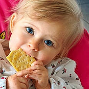 Mya a rejoint le concours — aidez-le/la à gagner de superbes lots ! baby, blue_eyes, child, close_up, cracker, cute, eating, face, floral_pattern, hands, indoor, light_blond_hair, person, pink_background, portrait, seated, snack, toddler, white_clothing, young_child