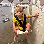 Jayden is registered to the contest to win money with this photo: toddler, child, bathroom, child_seat, yellow_onesie, straps, tiles, curious, hospital_wristband, barefoot, floor_tiles, handrail, wall, indoor, person, young_child, sitting, safety_strap, expression, plaster