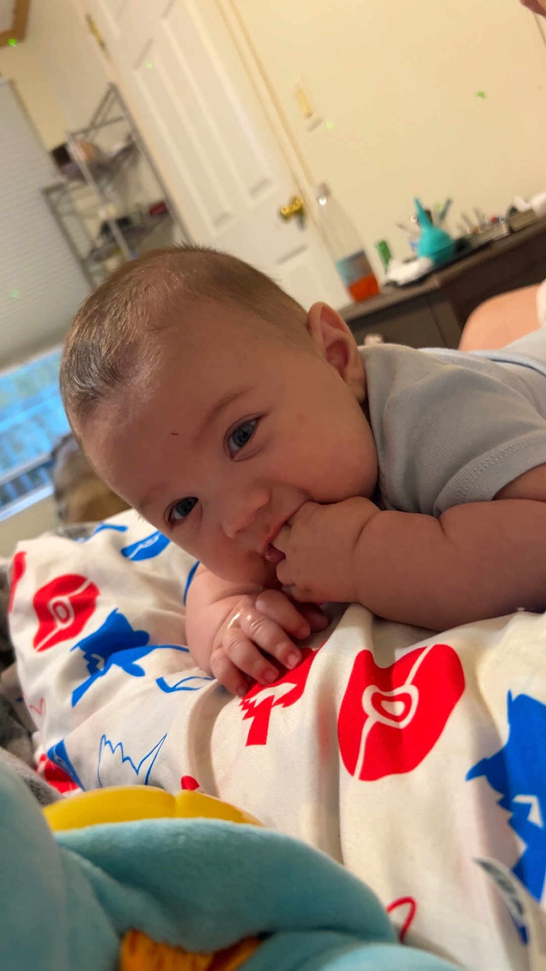 Zayden is registered to the contest to win money with this photo: baby, infant, child, blanket, hand, face, fingers, toy, indoor, curious, lying_down, blue_eyes, short_hair, soft_toy, colorful, patterned_blanket, door, shelf, room, closeup