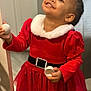 toddler, child, smiling, red_dress, holiday_outfit, indoor, holding_object, door_frame, happy, person, young_child, festive_clothing, cute, portrait, celebration, girl, bright_colors, face, looking_up, candid