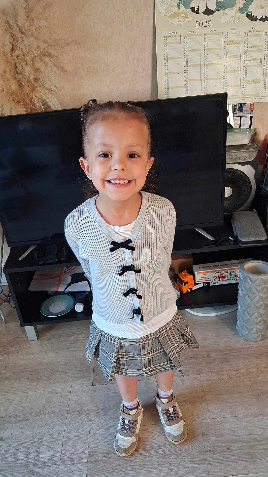 Charlie participe au concours pour gagner de l'argent avec cette photo : child, girl, smiling, standing, grey_sweater, pleated_skirt, sneakers, television, calendar_2026, toy_truck, shelf, speaker, plate, wood_floor, indoor, happy, braided_hair, black_bow, living_room, portrait