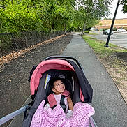 Zoey is registered to the contest to win money with this photo: baby, car_seat, stroller, blanket, pink, sidewalk, trees, greenery, parking_lot, outdoor, happy, infant, person, nature, daylight, road, urban, child, walk, leaves