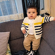 Amir participe au concours pour gagner de l'argent avec cette photo : cardigan, casual_clothing, child, couch, cozy, curtains, expression, face, furniture, home, indoor, pants, person, pillow, sneakers, standing, striped_clothing, toddler, window, young_child