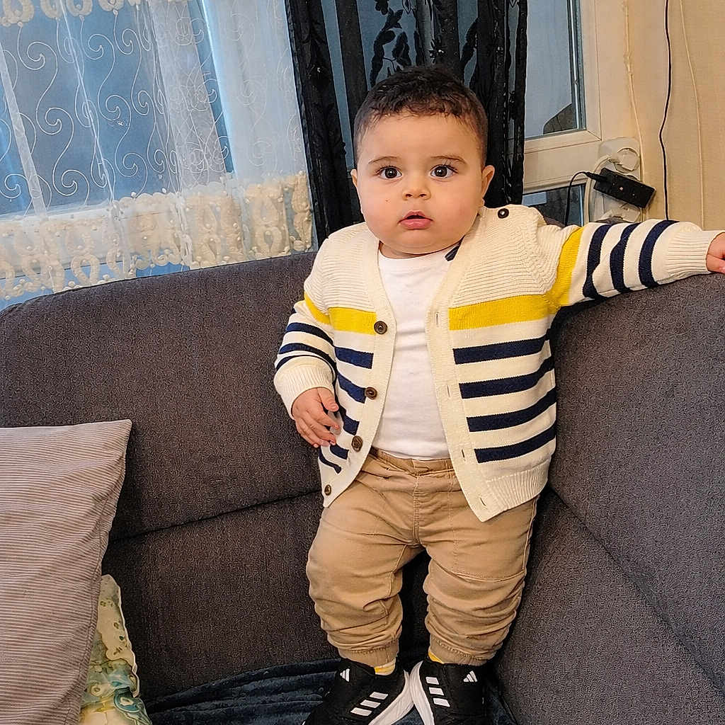 Amir participe au concours pour gagner de l'argent avec cette photo : cardigan, casual_clothing, child, couch, cozy, curtains, expression, face, furniture, home, indoor, pants, person, pillow, sneakers, standing, striped_clothing, toddler, window, young_child