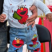 Tyce is registered to the contest to win money with this photo: toddler, child, smiling, happy, standing, jeans, sneakers, elmo, cartoon, clothing, person, hand, indoor, chair, denim, cute, curly_hair, white_shirt, blue_jeans, footwear