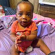 Quinton is registered to the contest to win money with this photo: baby, infant, child, clapping, purple_sheet, pacifier, bed, wooden_wall, air_conditioner, indoors, pink_blanket, orange_onesie, person, cute, sitting, face, expression, hair_bun, room, furniture