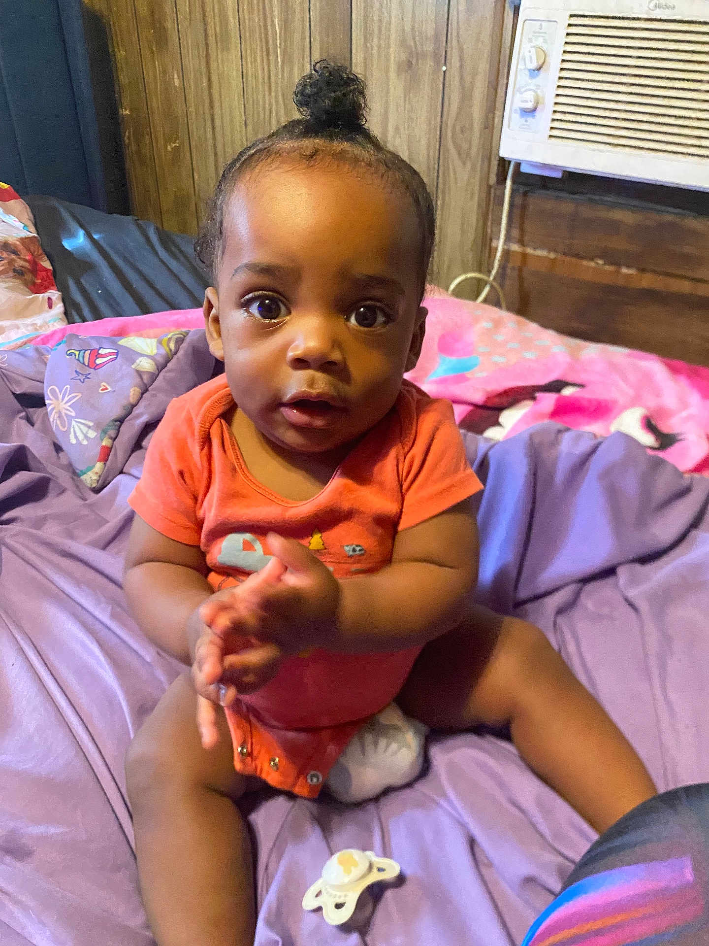 Quinton is registered to the contest to win money with this photo: baby, infant, child, clapping, purple_sheet, pacifier, bed, wooden_wall, air_conditioner, indoors, pink_blanket, orange_onesie, person, cute, sitting, face, expression, hair_bun, room, furniture