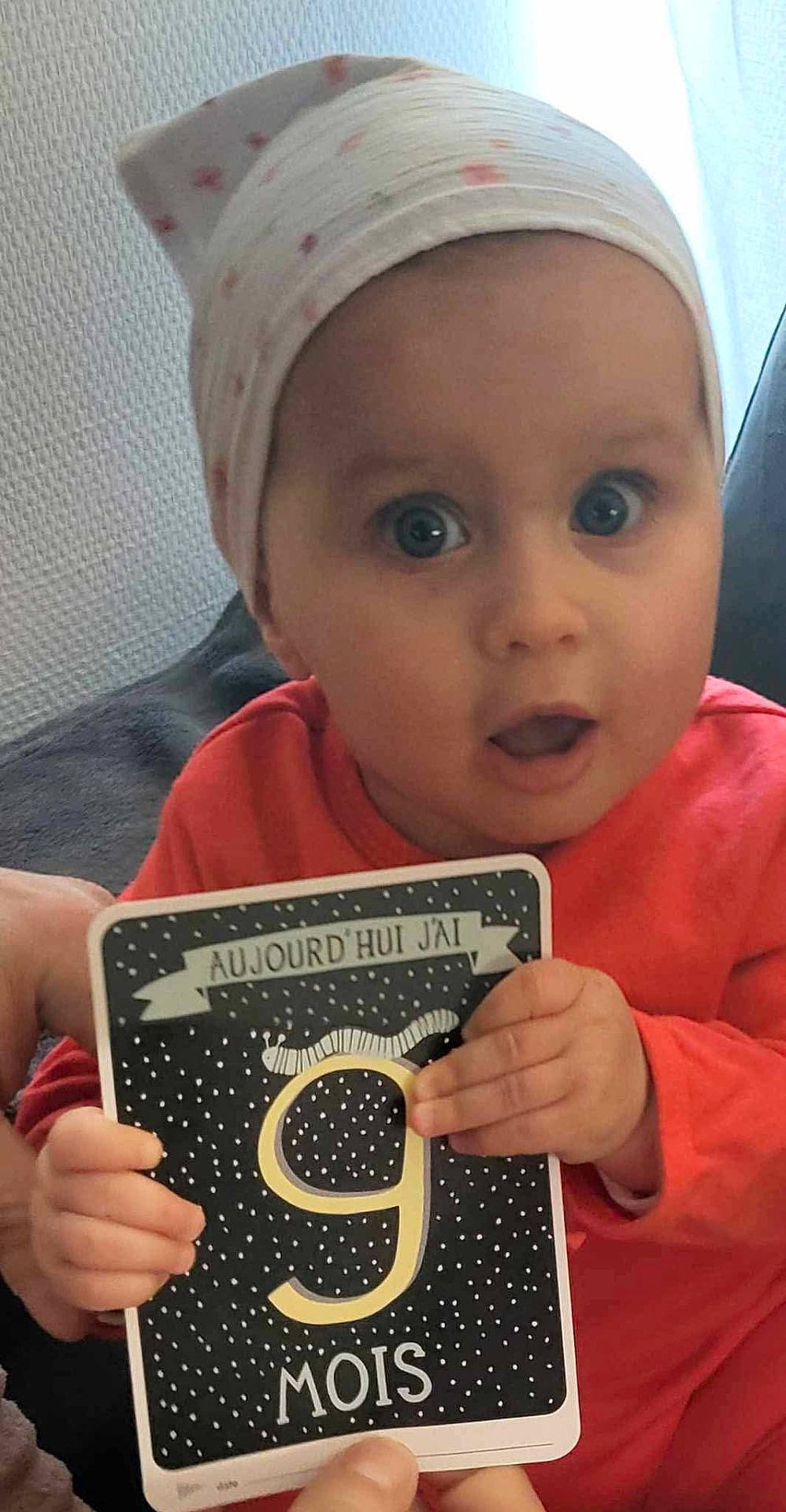 Thaïs participe au concours pour gagner de l'argent avec cette photo : accessories, baby, bodypart, cap, clothing, couch, creditcard, face, finger, furniture, hand, hat, head, number, person, photography, portrait, symbol, t_shirt, text