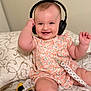 baby, headphones, smiling, floral_dress, plush_toy, bed, blanket, infant, cute, happy, indoors, child, person, sitting, toy, playful, candid, soft_lighting, closeup, expression
