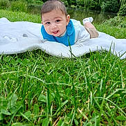 Ayden a rejoint le concours — aidez-le/la à gagner de superbes lots ! baby, blanket, blue_bib, child, clothing, cute, daylight, face, grass, greenery, happy, head, infant, lying_down, nature, outdoor, pond, portrait, smiling, trees