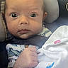 baby, infant, car_seat, blanket, clothing, face, head, hand, sitting, wide_eyes, curious, indoor, safety_label, seat_belt, newborn, child, person, portrait, cute, cozy
