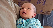 Eleanor joined the competition — help win amazing prizes! baby, infant, person, clothing, onesie, holding, arm, cardboard_box, indoor, bed, bedroom, technology, router, blue, patterned_clothing, face, skin, newborn, cute, resting