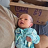 baby, infant, person, clothing, onesie, holding, arm, cardboard_box, indoor, bed, bedroom, technology, router, blue, patterned_clothing, face, skin, newborn, cute, resting