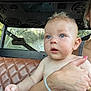 baby, child, person, hand, arm, skin, blue_eyes, hair, diaper, seat, leather, car_interior, window, sunlight, adult, bracelet, nail_polish, vehicle, curious, holding