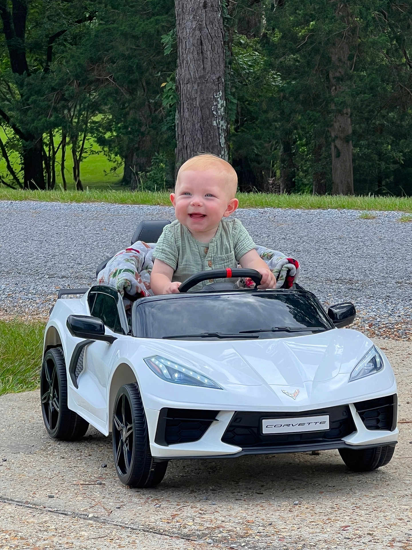 Jon Lewis joined the competition — help win amazing prizes! baby, child, toy_car, corvette, white_car, outdoor, smiling, happy, steering_wheel, blanket, grass, trees, road, toy_vehicle, small_car, infant, daylight, nature, cute, playing