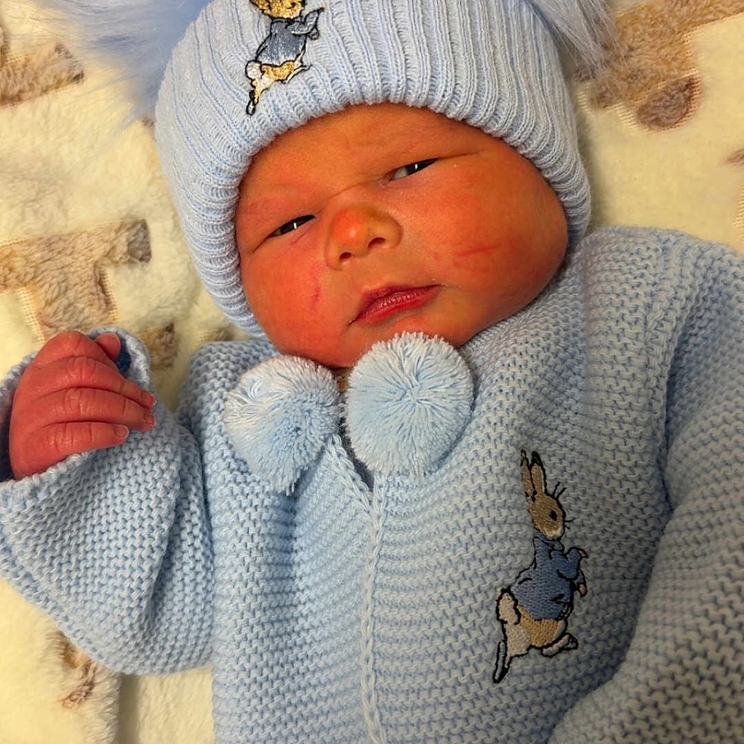 Frankie-James is registered to the contest to win money with this photo: baby, blanket, blue_clothing, child, cozy, cute, face, hand, hat, indoor, infant, knitwear, newborn, pom_poms, portrait, resting, sleepy, smile, soft_texture, warm_clothing