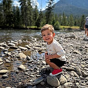 Liam is registered to the contest to win money with this photo: child, toddler, smiling, river, rocks, riverbank, outdoor, nature, mountains, pine_trees, bokeh, portrait, playful, squatting, red_shoes, tshirt, shorts, sunny, pebbles, vacation