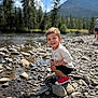 child, toddler, smiling, river, rocks, riverbank, outdoor, nature, mountains, pine_trees, bokeh, portrait, playful, squatting, red_shoes, tshirt, shorts, sunny, pebbles, vacation
