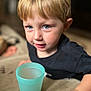 child, toddler, blond_hair, blue_eyes, portrait, close_up, indoor, plastic_cup, drink, hand, couch, shallow_depth_of_field, casual_clothing, cute, curious, smiling, gaze, domestic, young_boy, bokeh