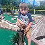 artificial_turf, boy, brick_border, child, clouds, crocs, fence, greenery, miniature_golf_course, ornamental_grass, outdoor, potted_plant, putter, rock, serious_expression, shorts, sky, t_shirt, wooden_deck, wooden_post