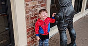 Grayson is registered to the contest to win money with this photo: child, boy, statue, brick_wall, sidewalk, building, window, spiderman_hoodie, smiling, outdoor, casual_clothing, footwear, toy_shoes, playful, happy, daylight, urban, posing, person, sculpture