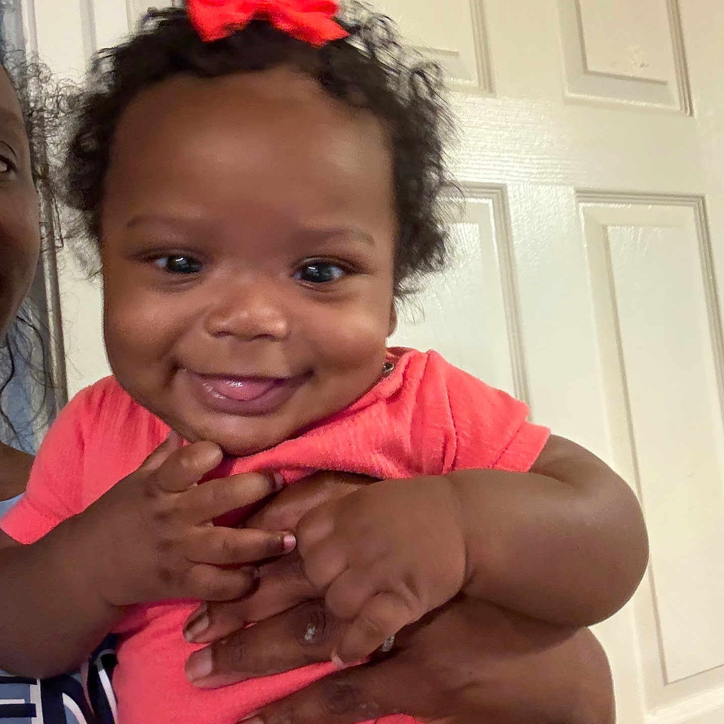 Curcedes is registered to the contest to win money with this photo: adult_hand, baby, child, closeup, clothing, curly_hair, cute, door, face, hand, happy, holding, indoor, infant, person, pink_shirt, portrait, red_bow, skin, smiling